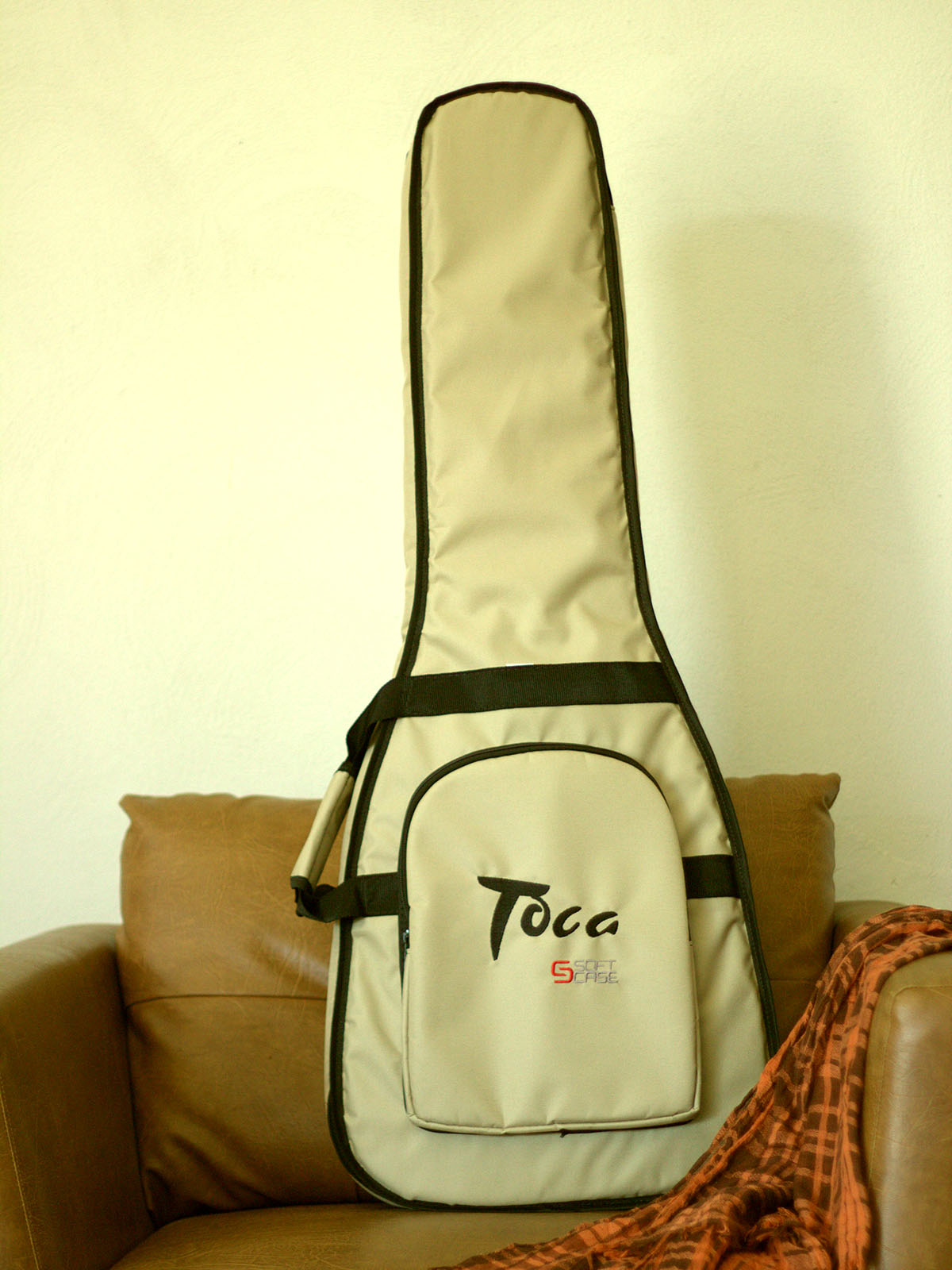 Capa para violão em tom bege com acabamentos pretos, apoiada em uma poltrona de couro